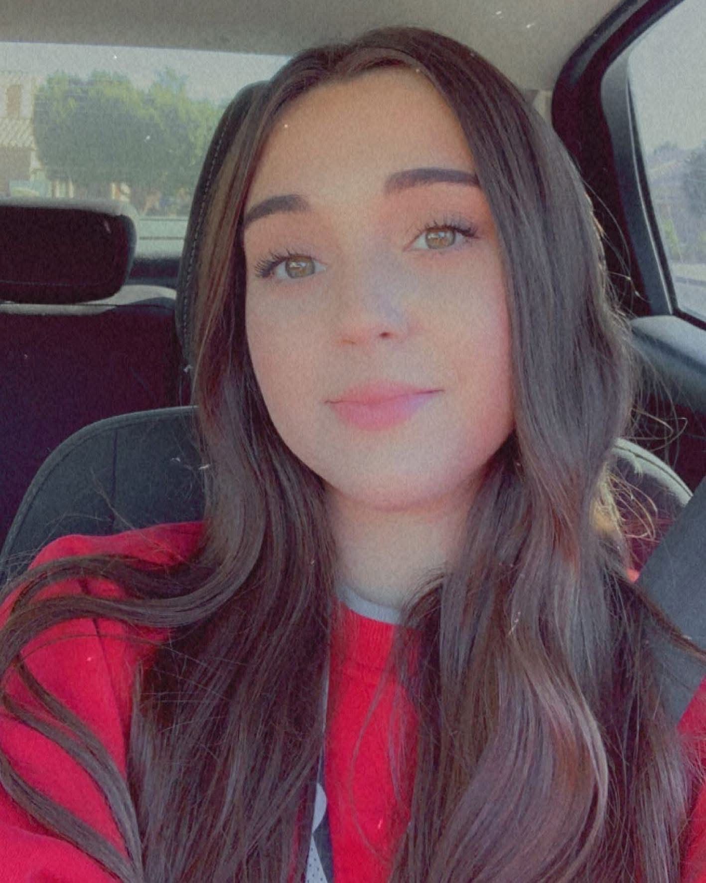 Woman with long brown hair in a red sweater smiles, sitting in a car with a seatbelt on.