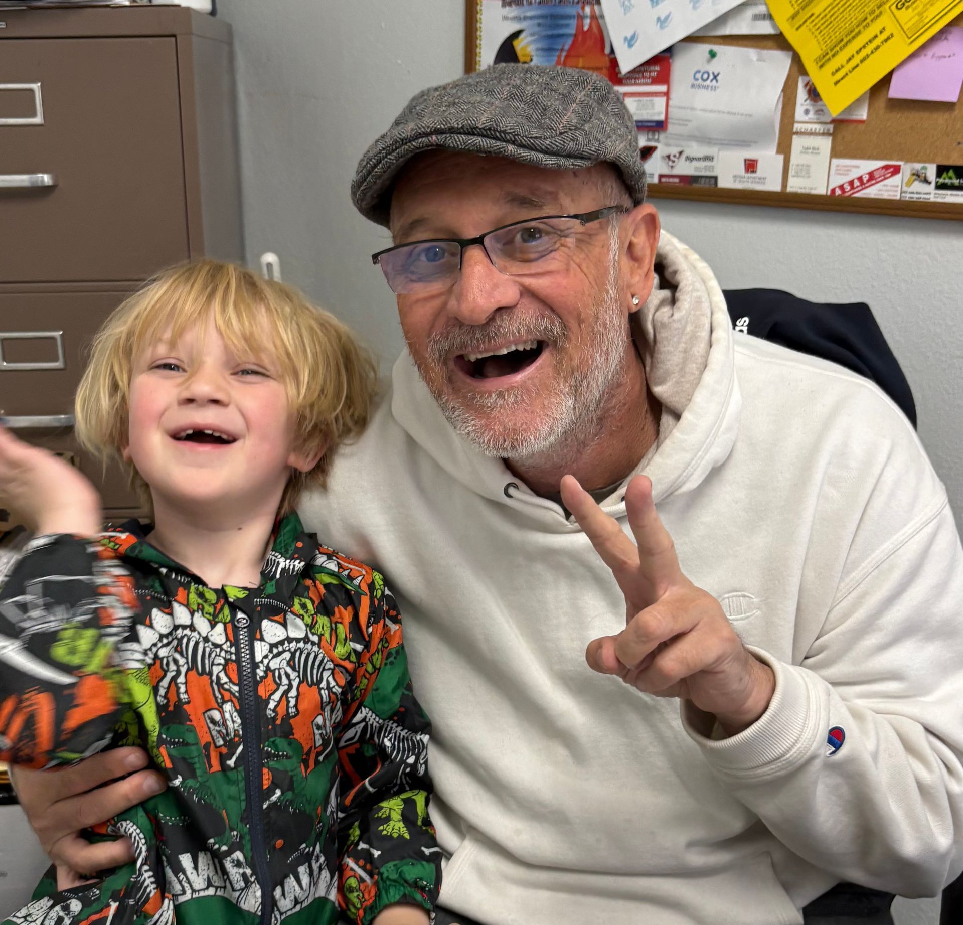Man with a grey cap and a child smiling at the camera. The man is holding up a peace sign. They are indoors.