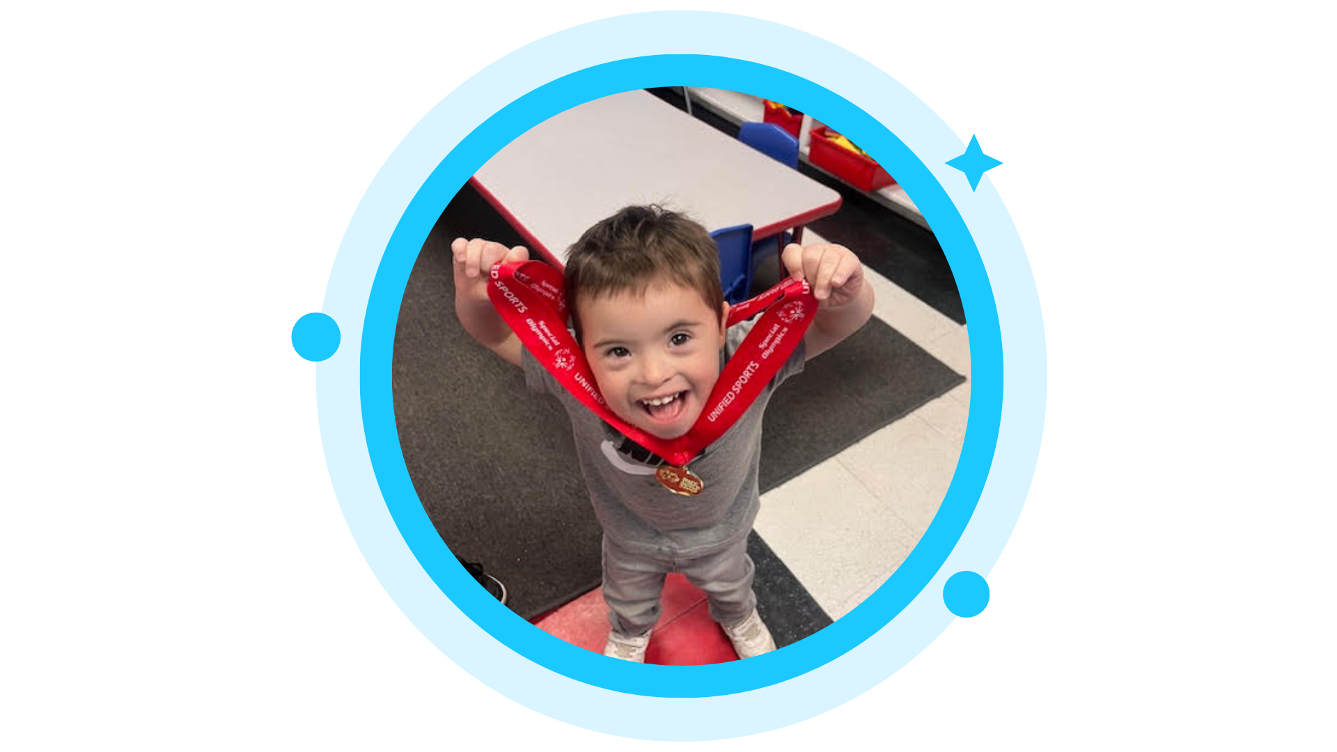 Boy smiles, holding up a red medal in a classroom.