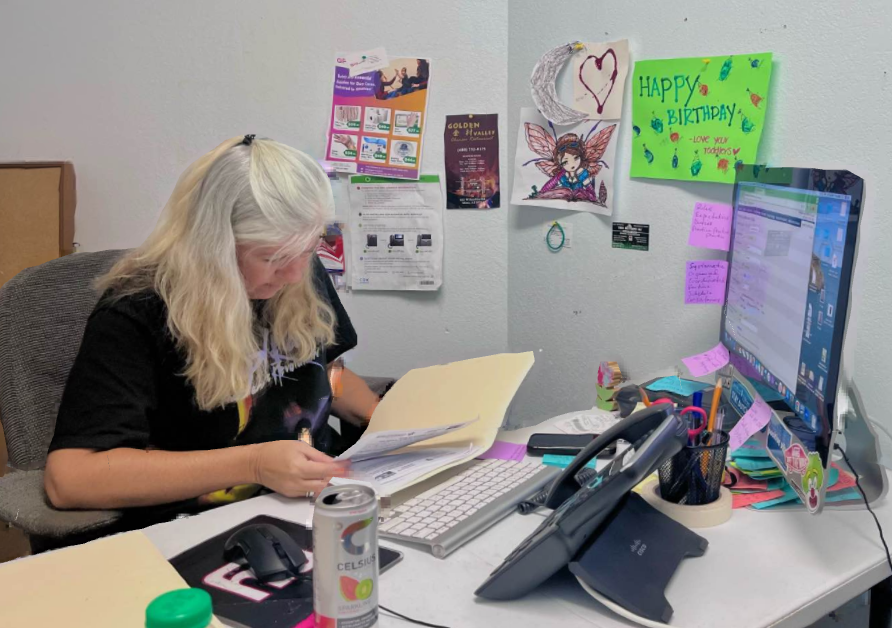 Woman with gray hair seated at a desk, looking at paperwork. Office setting with computer, phone, and decorations.