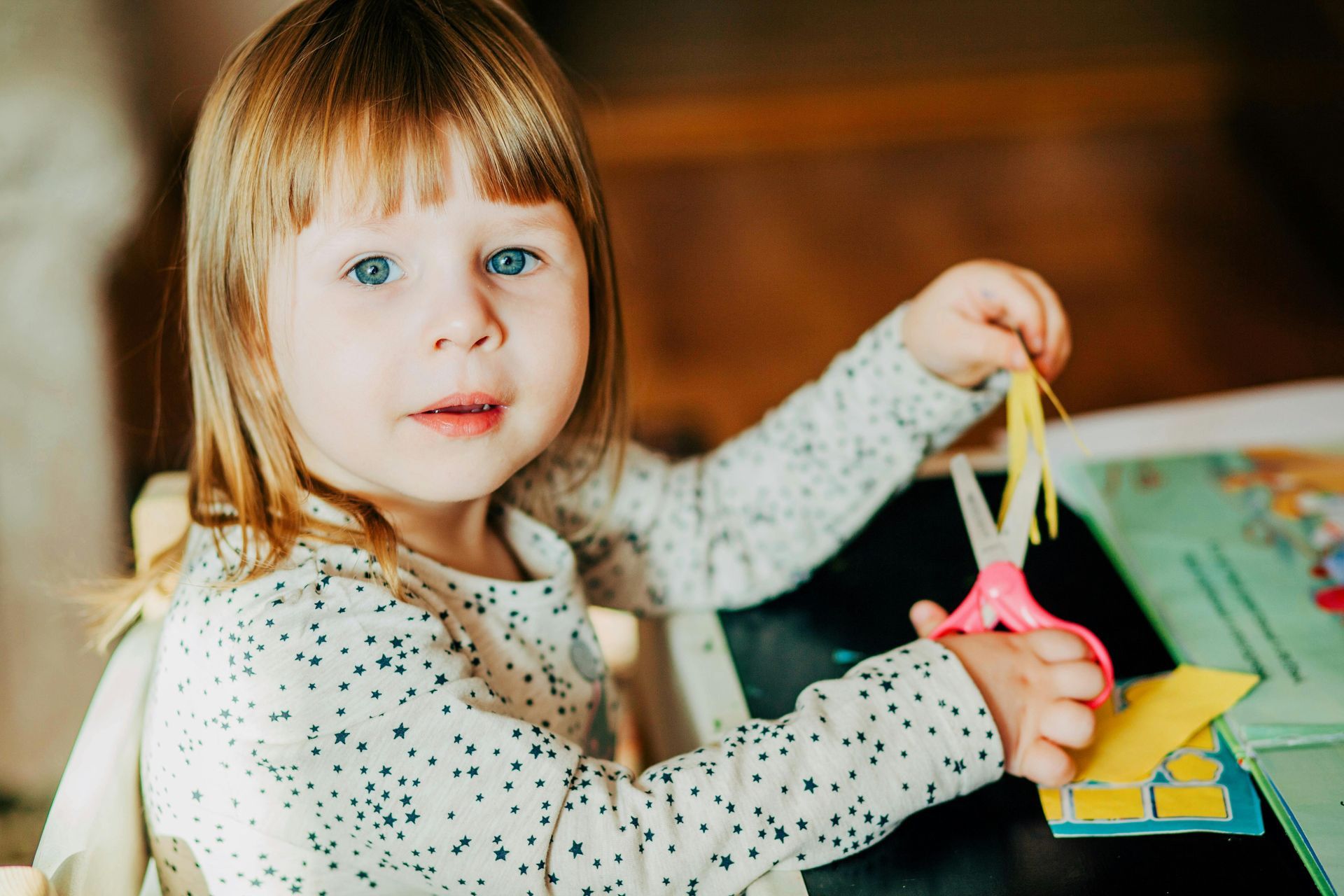 Young child with blue eyes cuts yellow paper with pink scissors.