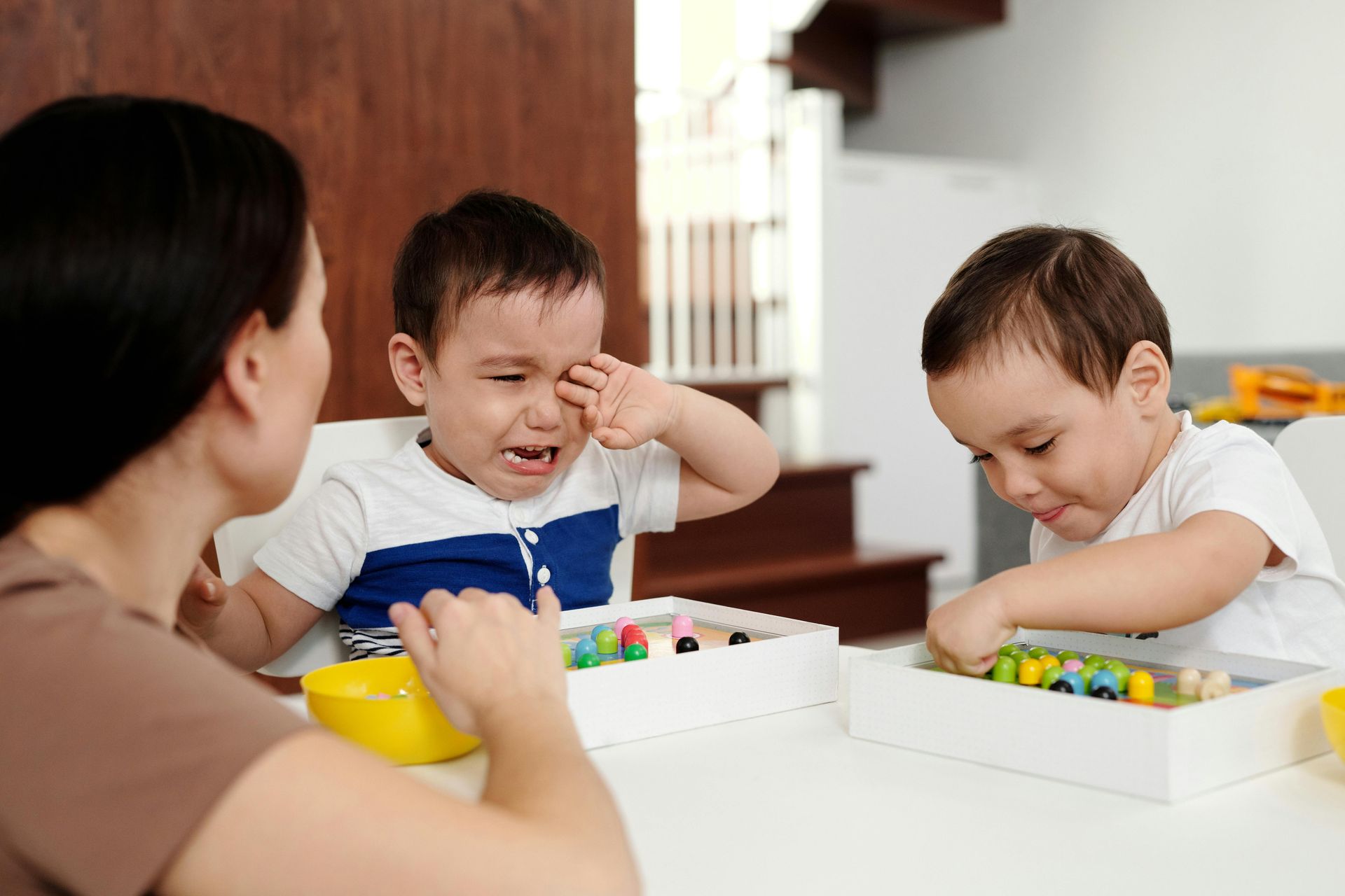 A child cries while another plays with toys at a table; adult sits nearby.