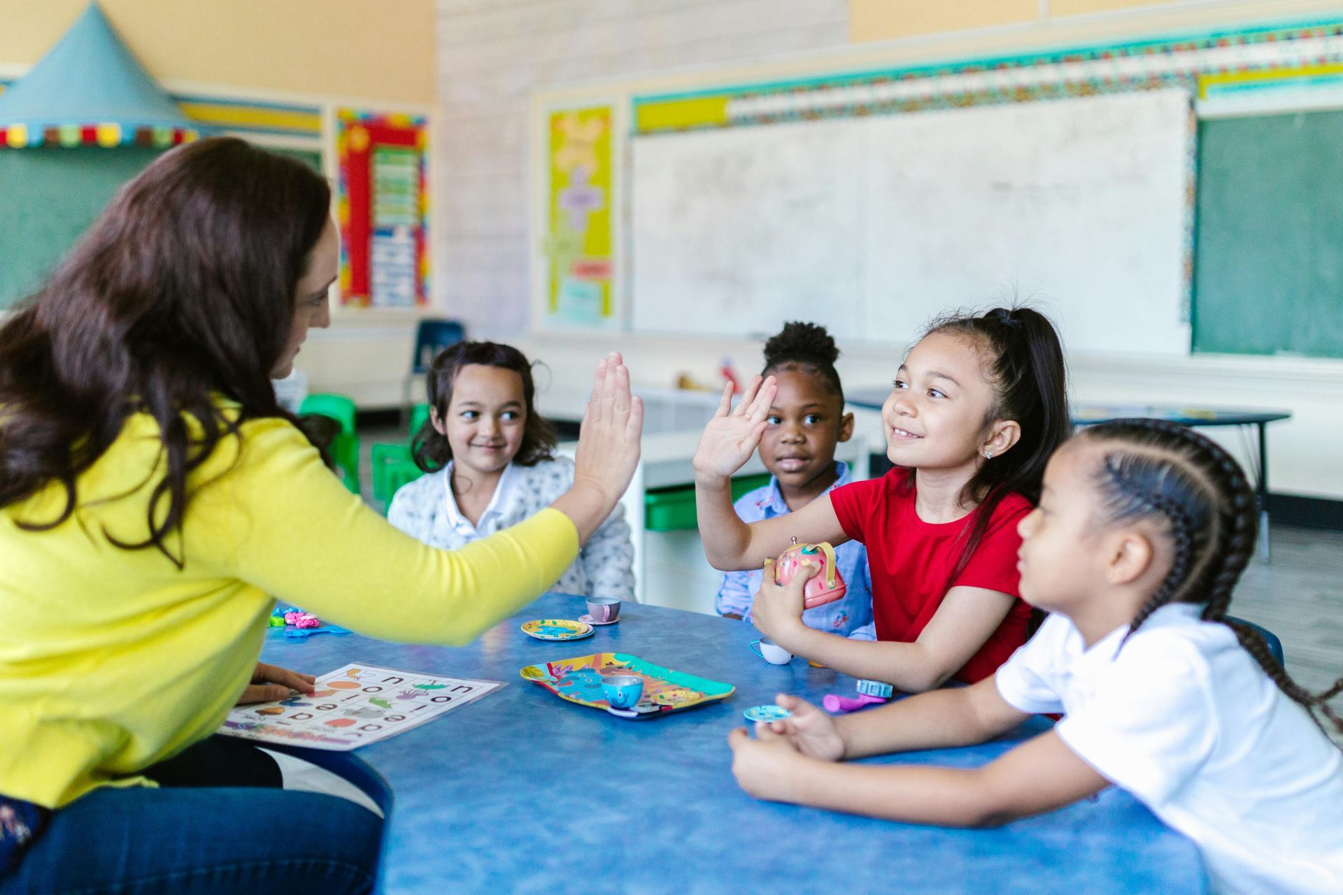 A teacher sits at a table in a classroom giving a high-five to a student, surrounded by other children playing.