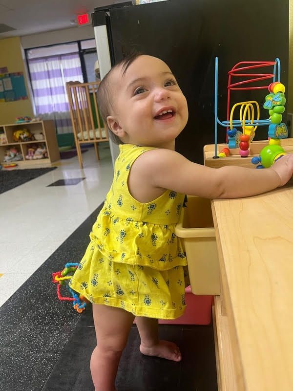 Smiling baby in a yellow dress stands at a wooden table, looking upward in a playroom.