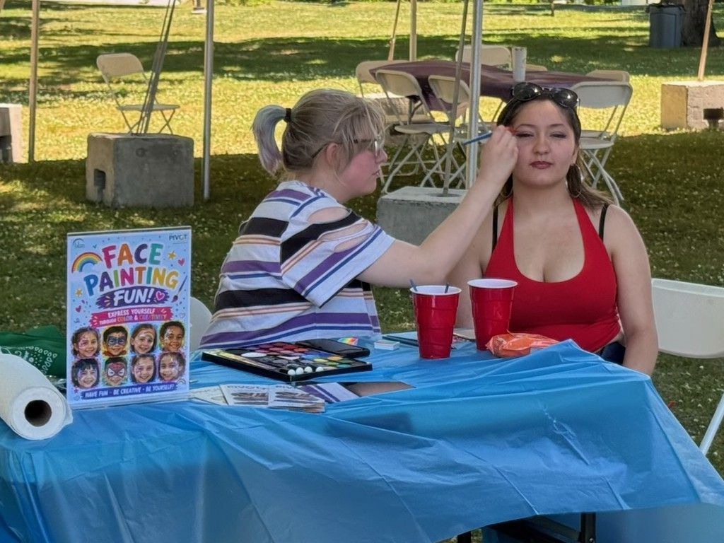 A woman does face painting to a girl sitting next to her. 
