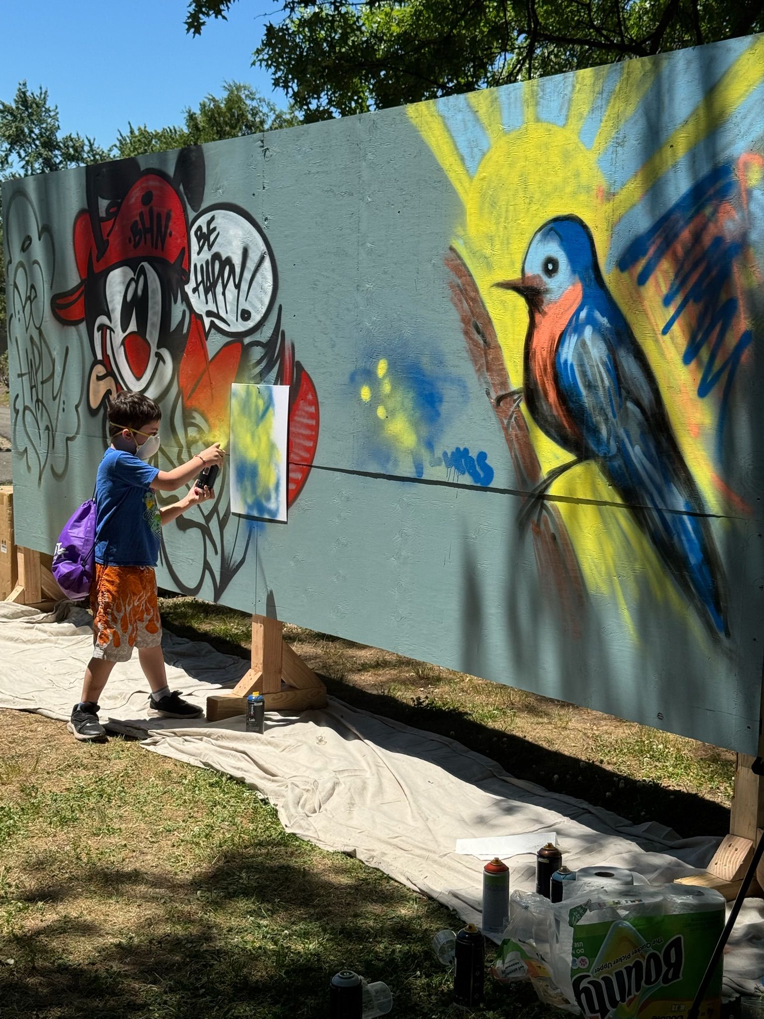 A child spray paints graffiti onto a large board that community members were spray painting together. The board featured a smiling cartoon character, a blue bird, and positive quotes like 