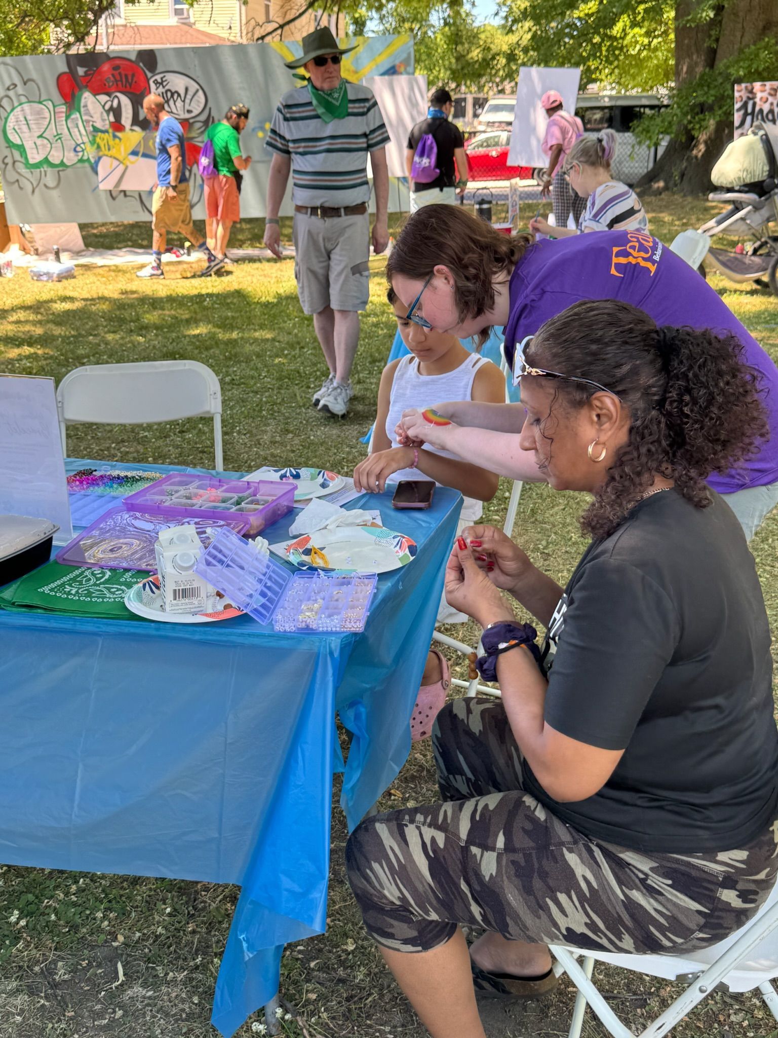 A woman sitting at a craft table making a beaded bracelet and a BHN staff member helping a young girl put on a newly created bracelet.
