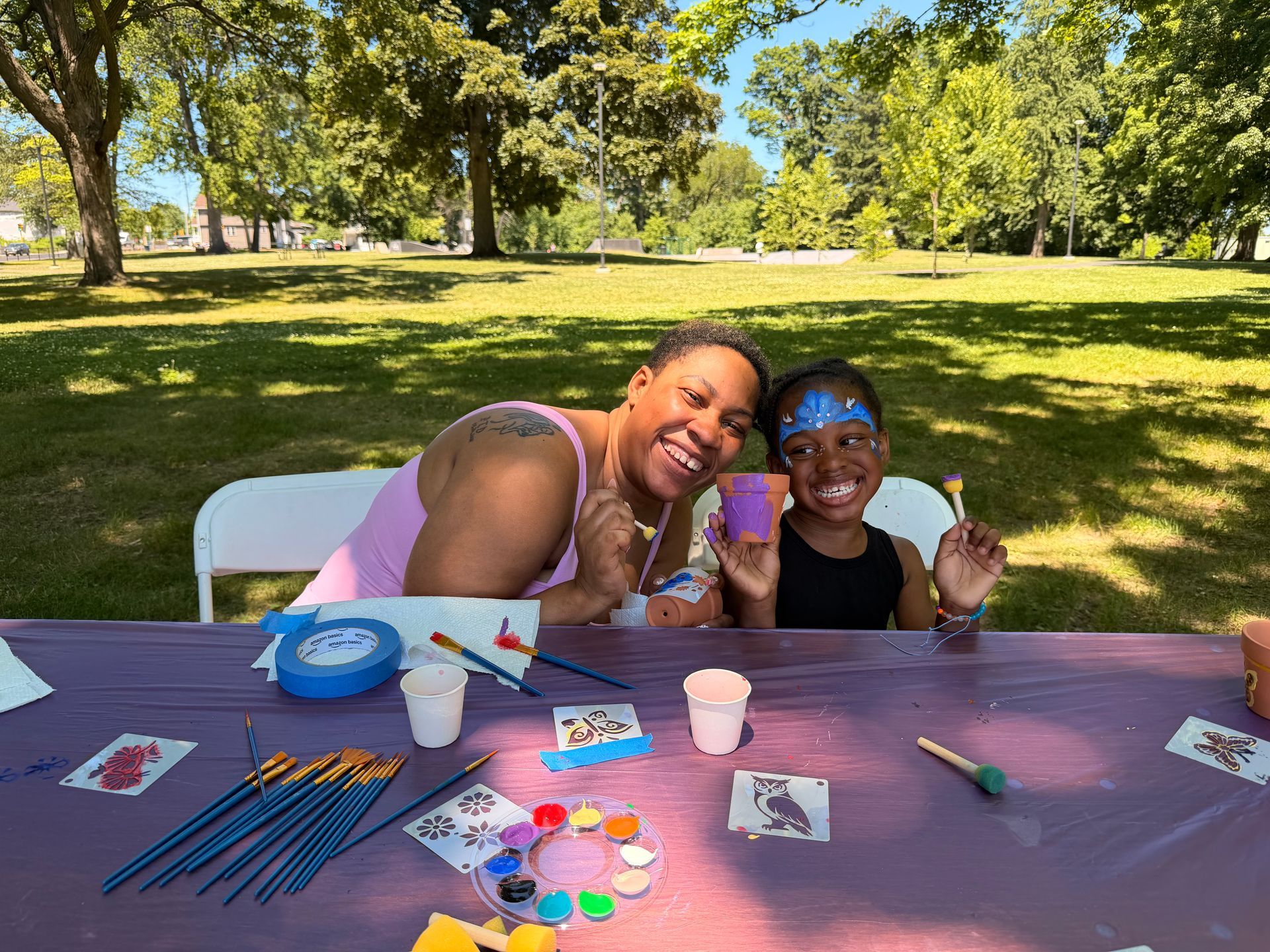 A woman and her young girl smile for a photo while painting terracotta pots in the park.