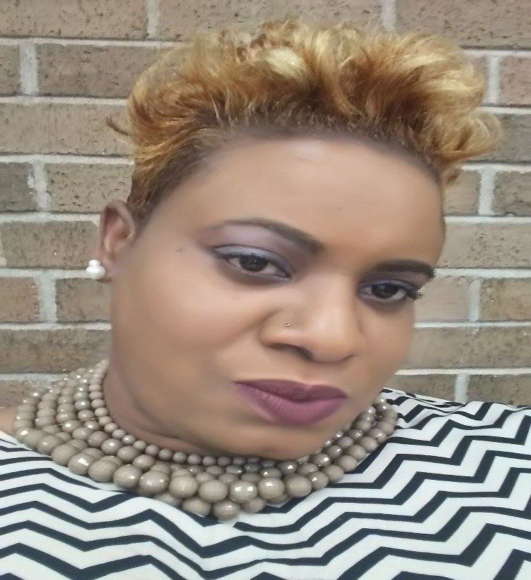 Close-up portrait of a woman with short blond hair, wearing a black-and-white zigzag top and pearl necklace, against a brick wall