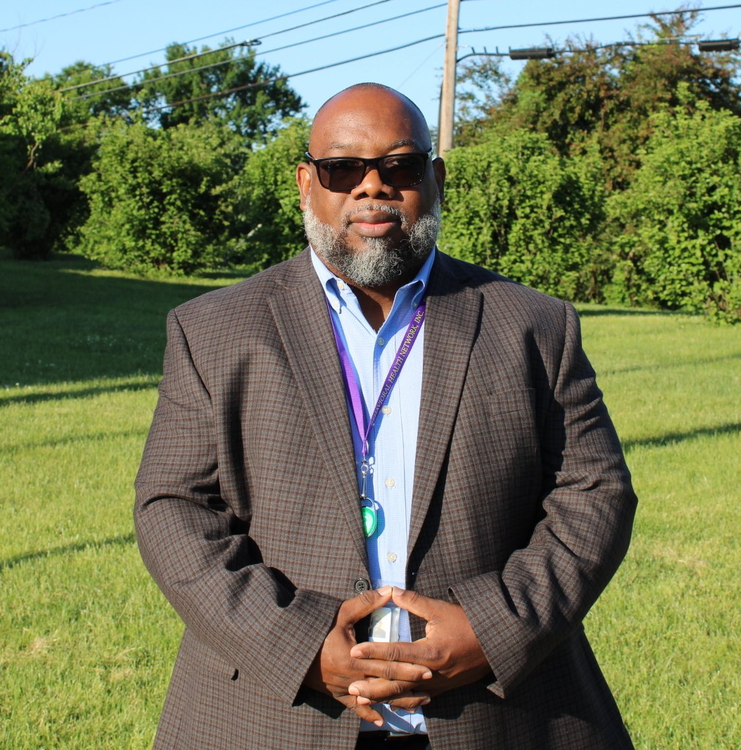 Man in sunglasses and brown blazer standing outdoors on grass in bright sunlight