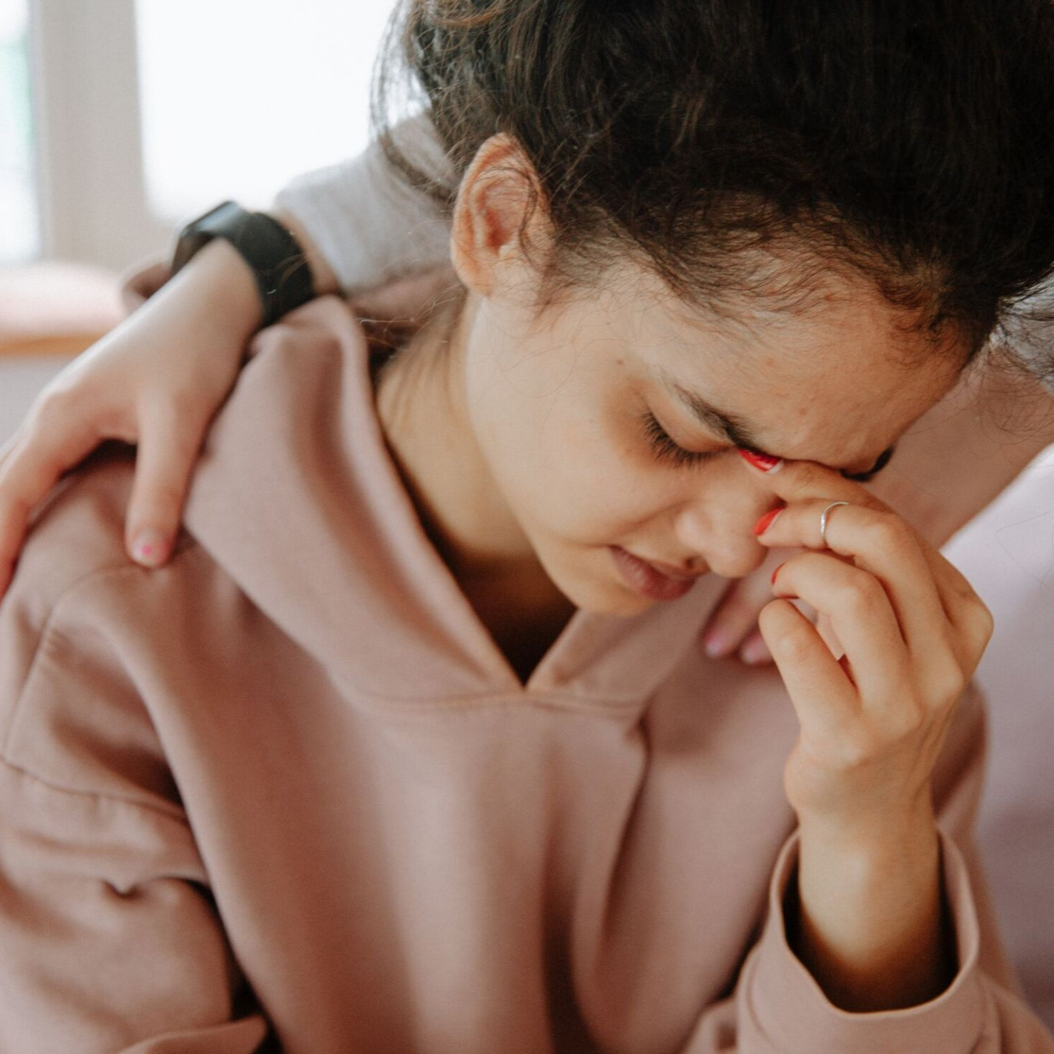 Woman in pink hoodie crying while another person comforts her with a hand on her shoulder