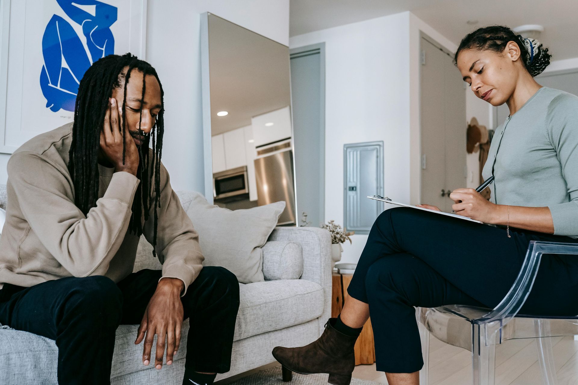 An image of a man sitting on a couch while speaking with a therapist. The man is leaning slightly forward while clasping his hands together. The therapist is taking notes.
