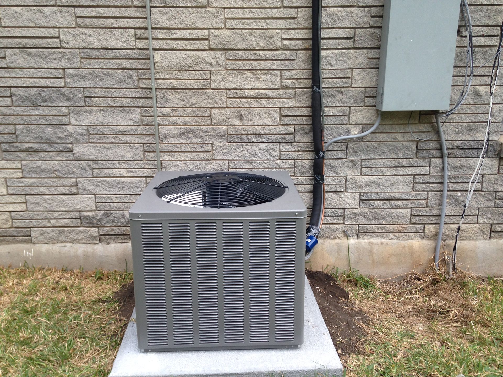 A gray air conditioner is sitting on a concrete base next to a brick wall.