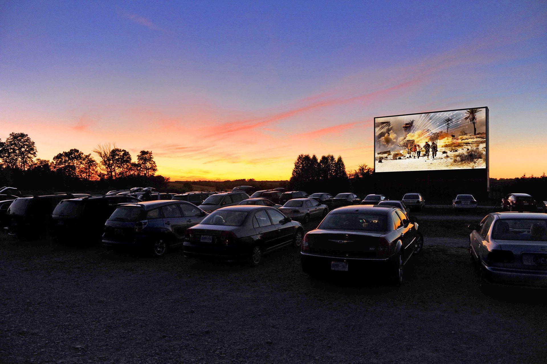 A lot of cars are parked in a drive in theater at sunset.