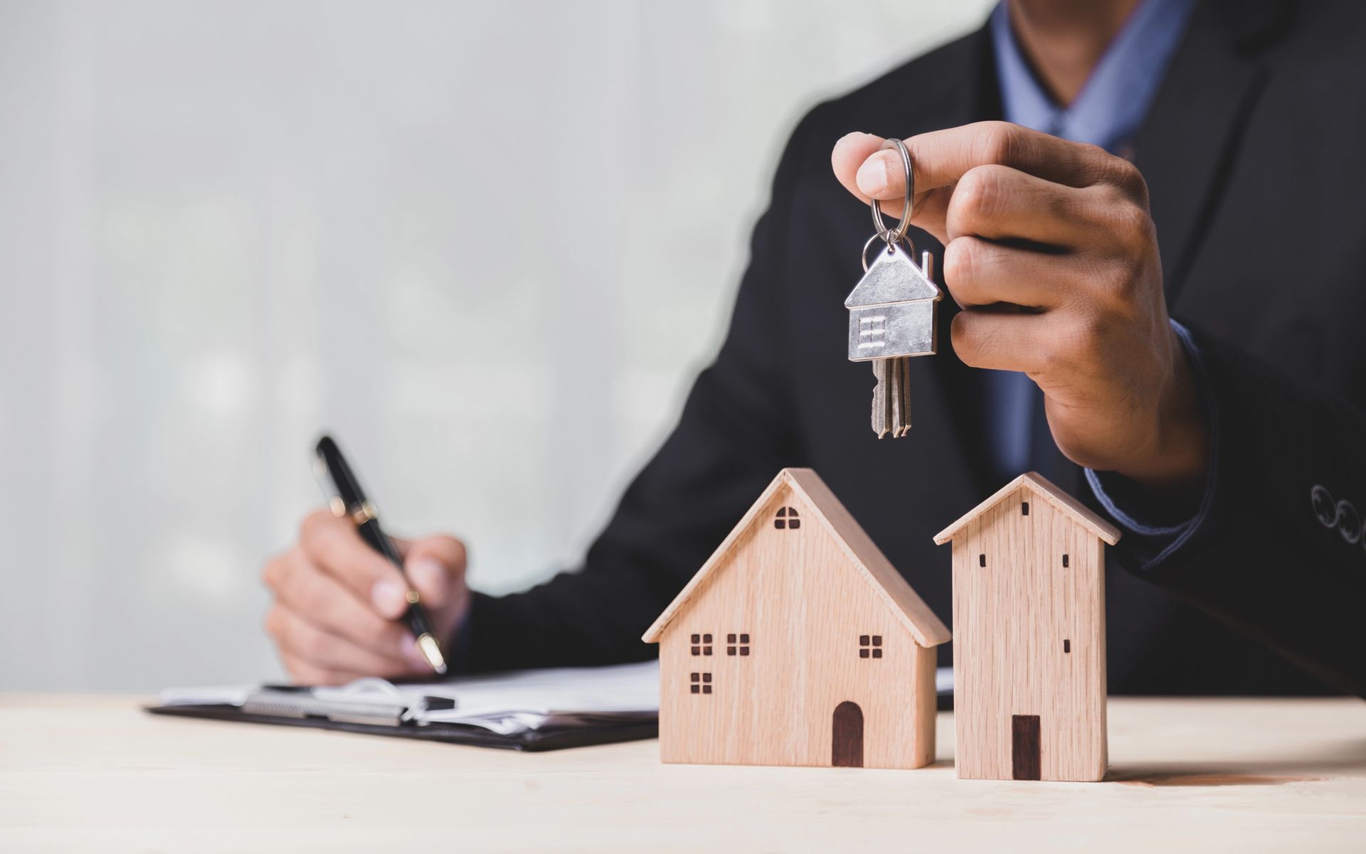 A real estate agent in a suit holding keys, with wooden house models on a desk and signing paperwork.
