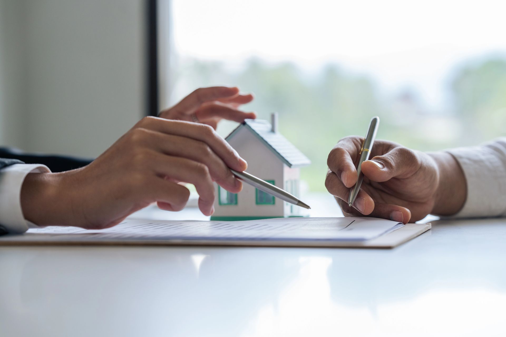 Hands placing a small model house on documents while another hand writes during a real estate meeting