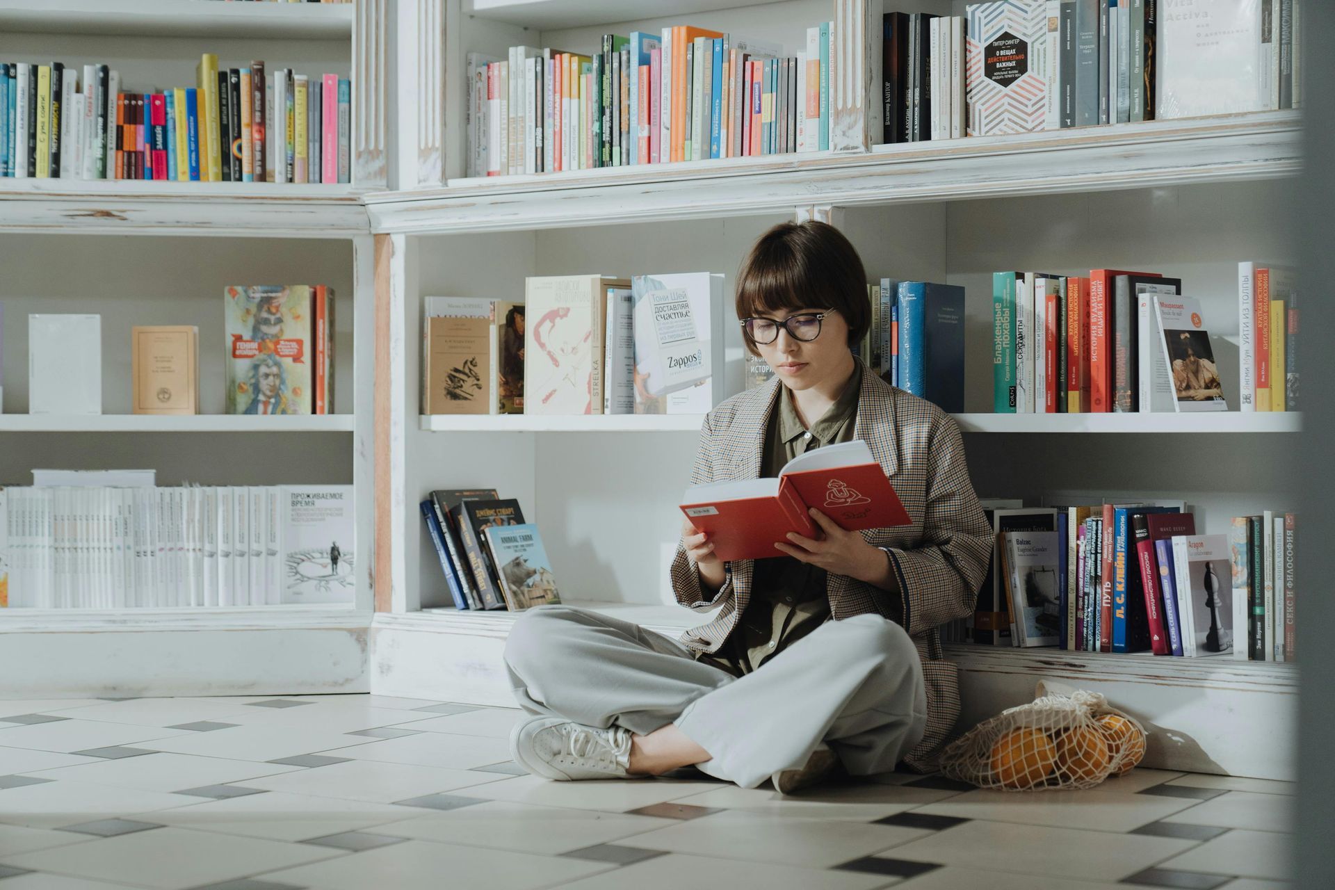 A woman is sitting on the floor reading a book in a library.