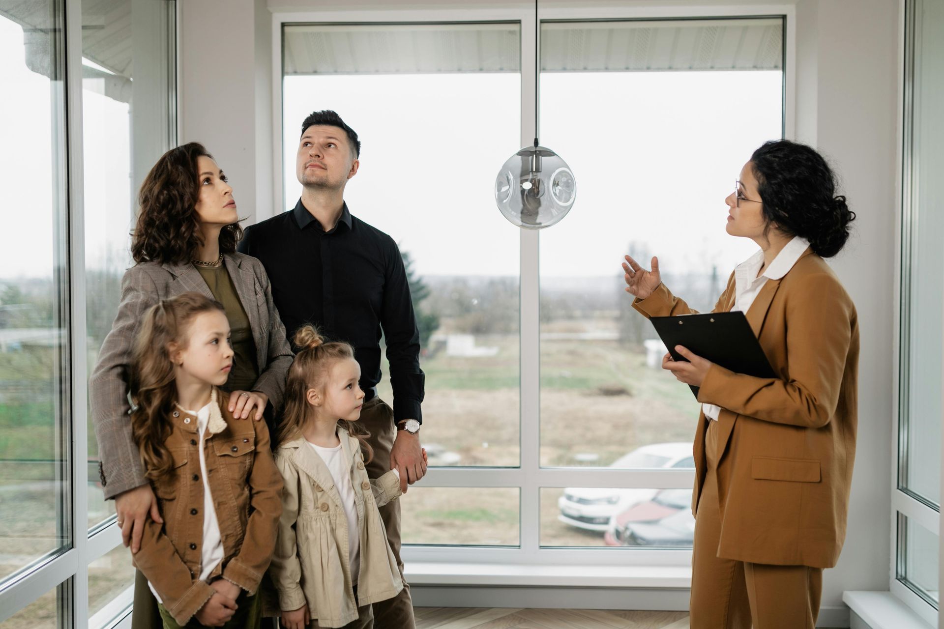A professional in a tan suit points to a window while showing a family through a bright, modern, unfurnished room.