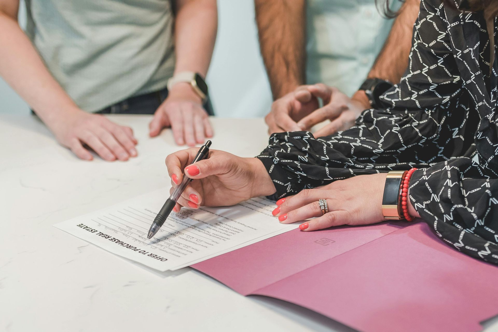 Person signing document with pen, other people's hands visible. Pink folder on white table.