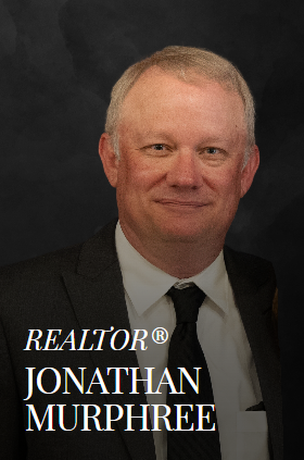 Jonathan Murphree, Realtor. Portrait of a man in a suit, smiling, with a black background.
