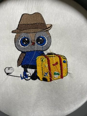 An embroidery of an owl wearing a brown hat and blue shirt, standing next to a yellow suitcase with travel stickers.