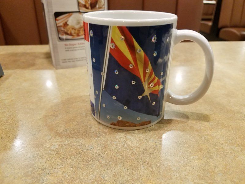 A white mug featuring a graphic of the Arizona state flag on a dark blue, star-speckled background, sitting on a table.
