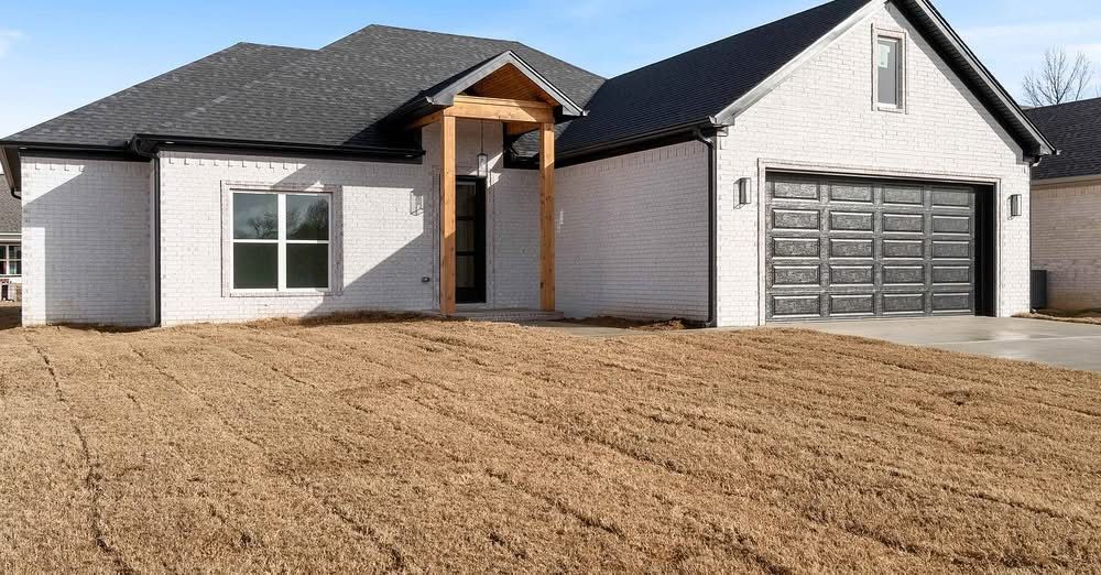 Single-story white brick house with a dark roof, wooden porch pillars, a two-car garage, and dry grass in the front yard.