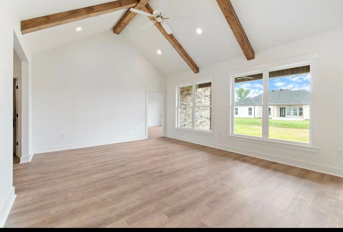 A spacious, unfurnished living room with vaulted ceilings, exposed wooden beams, white walls, and wood-look flooring.