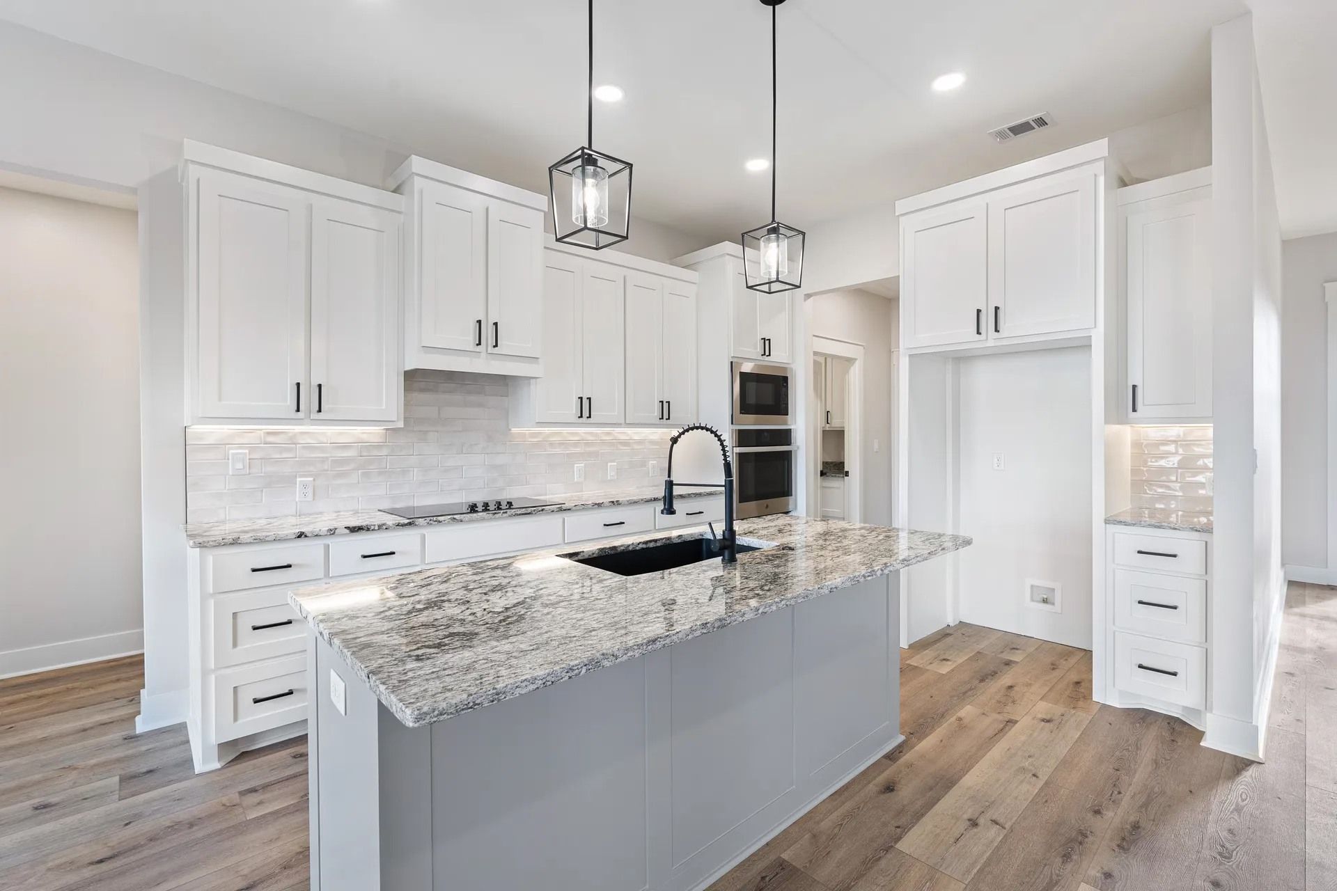A modern white kitchen with a gray island, speckled granite countertops, stainless appliances, and wood-look flooring.