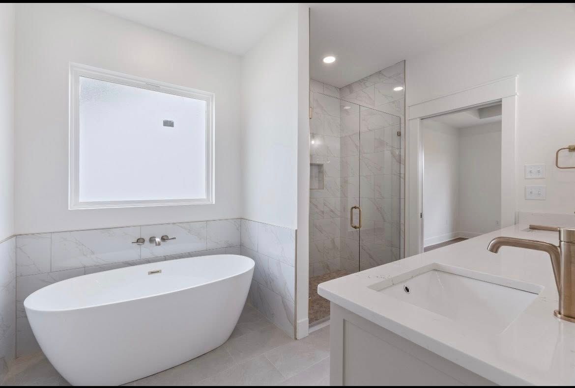 A bright bathroom with a freestanding white tub, a marble-tiled shower, and a sink with gold fixtures.