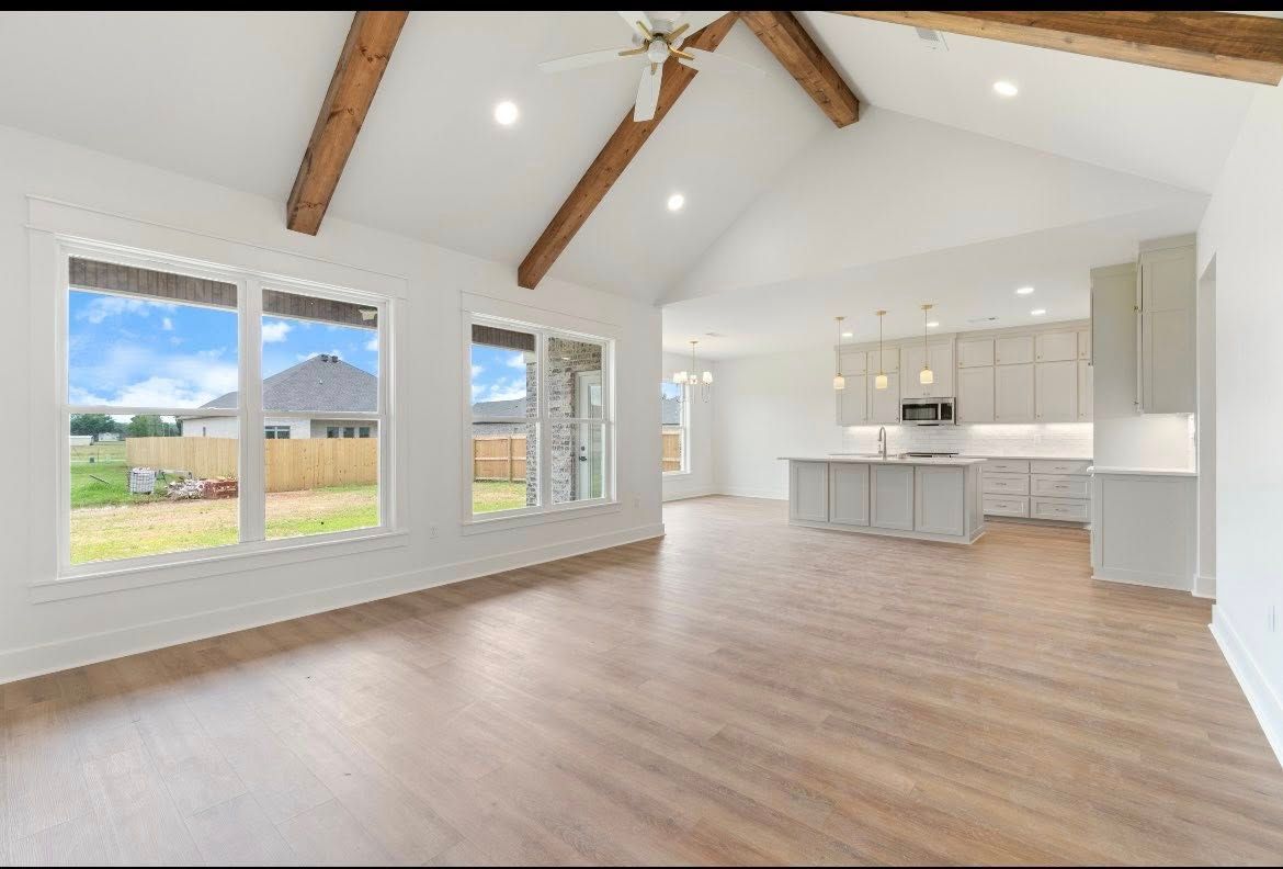 A bright, open-concept living area and kitchen with vaulted ceilings, exposed wooden beams, and light-toned flooring.