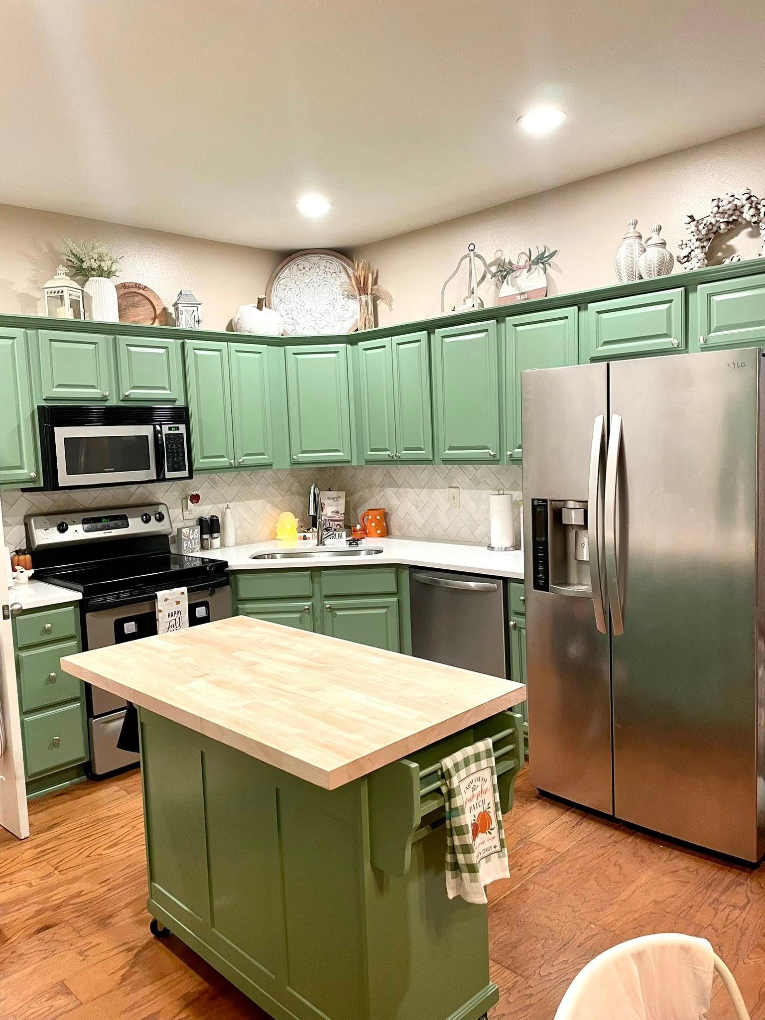 A kitchen with sage green cabinets, stainless steel appliances, a light-colored wooden island, and wood flooring.