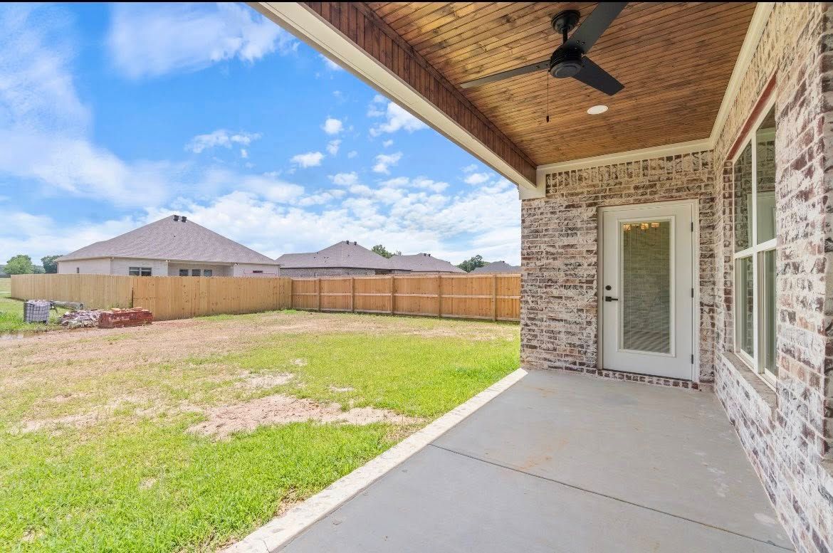 A covered concrete patio with a wooden ceiling and fan, overlooking a grassy backyard with a wooden fence.