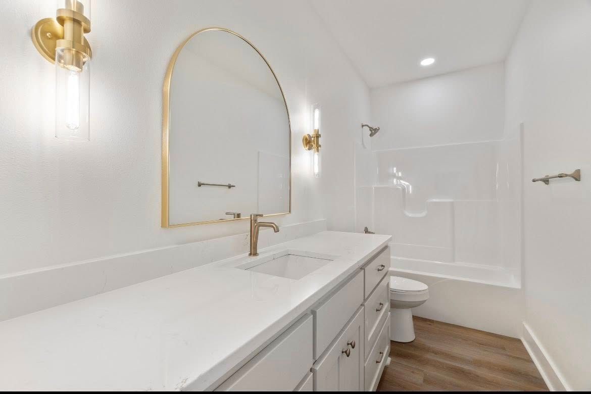 A modern white bathroom with a vanity, gold-rimmed arched mirror, gold sconces, and a shower-tub combo.