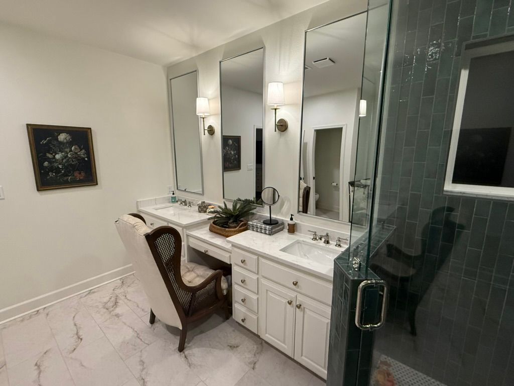 A bathroom vanity with white cabinets, two sinks, mirrors, and a decorative armchair, beside a tiled glass shower.
