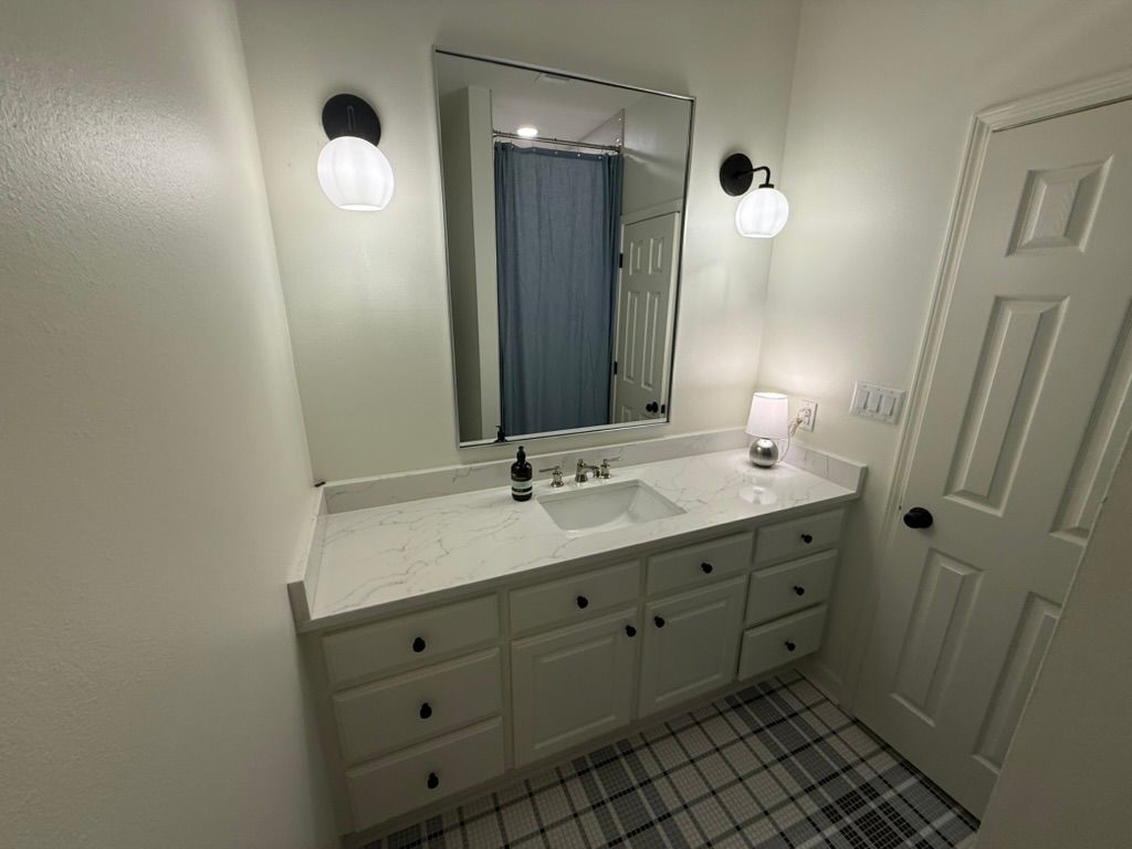 A white bathroom vanity with a mirror, two sconces, and a white sink, set against a room with checkered floor tiles.