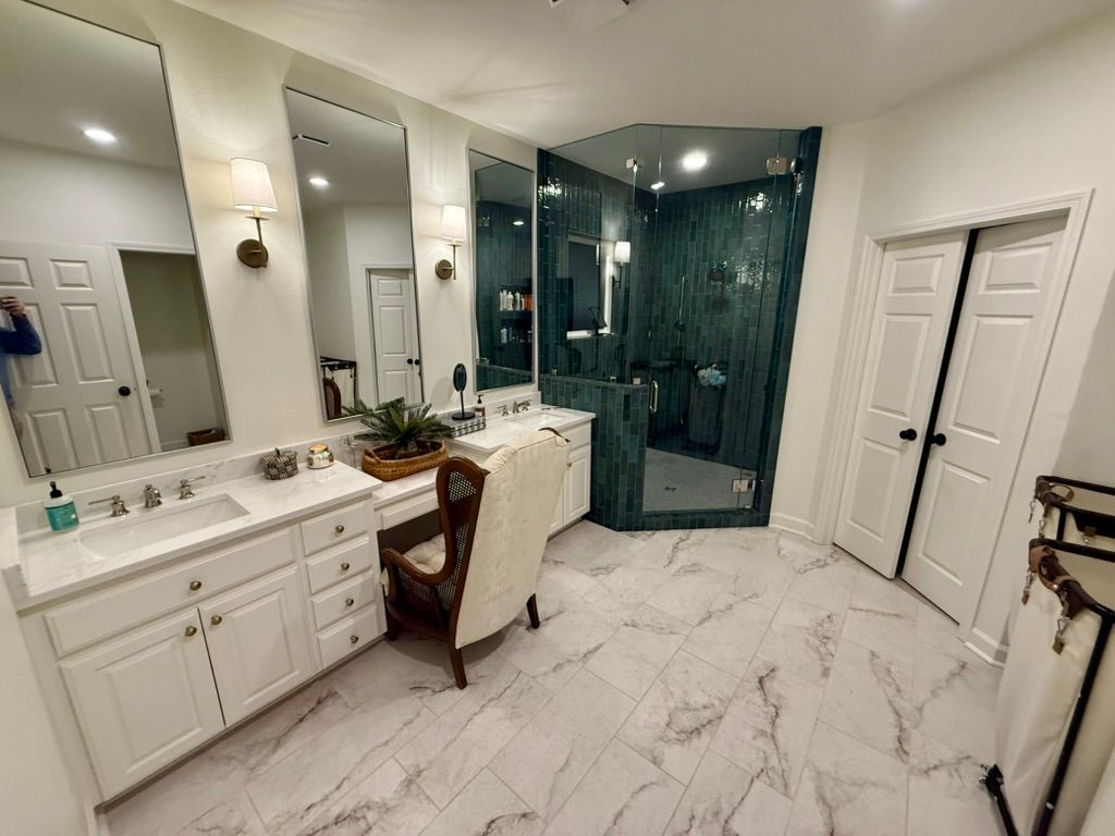 A bright bathroom with white vanity cabinets, dual mirrors, a makeup desk, and a large walk-in glass shower.