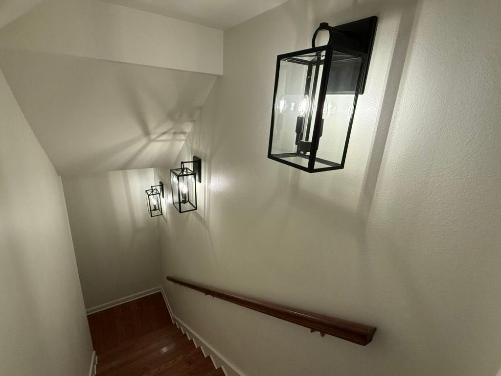 Wooden stairs lead down a hallway with beige walls, illuminated by three black metal lanterns and a wooden handrail.