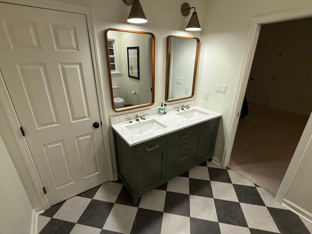 A bathroom with a dual-sink vanity, dark green cabinets, white countertops, two mirrors, and a black-and-white tiled floor.