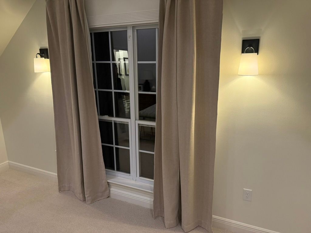 A window with beige curtains flanked by two wall-mounted lamps in a room with light-colored walls and carpet.