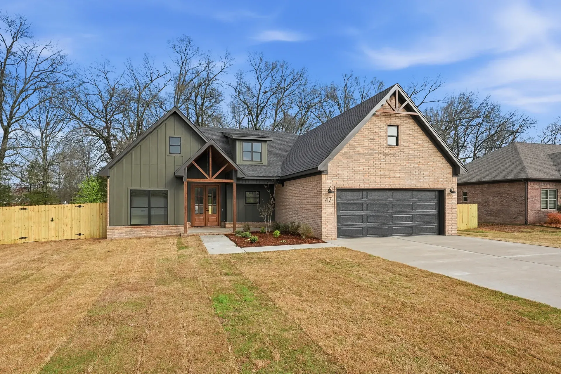 A modern house with a green exterior, brick facade, and garage, situated on a lawn with trees under a blue sky.