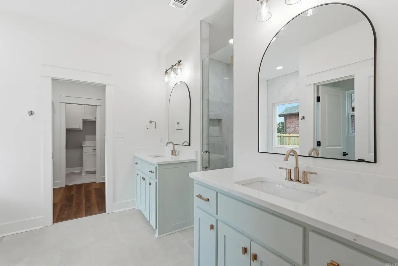 A bright bathroom featuring dual vanity cabinets in soft blue with gold hardware, white countertops, and arched mirrors.