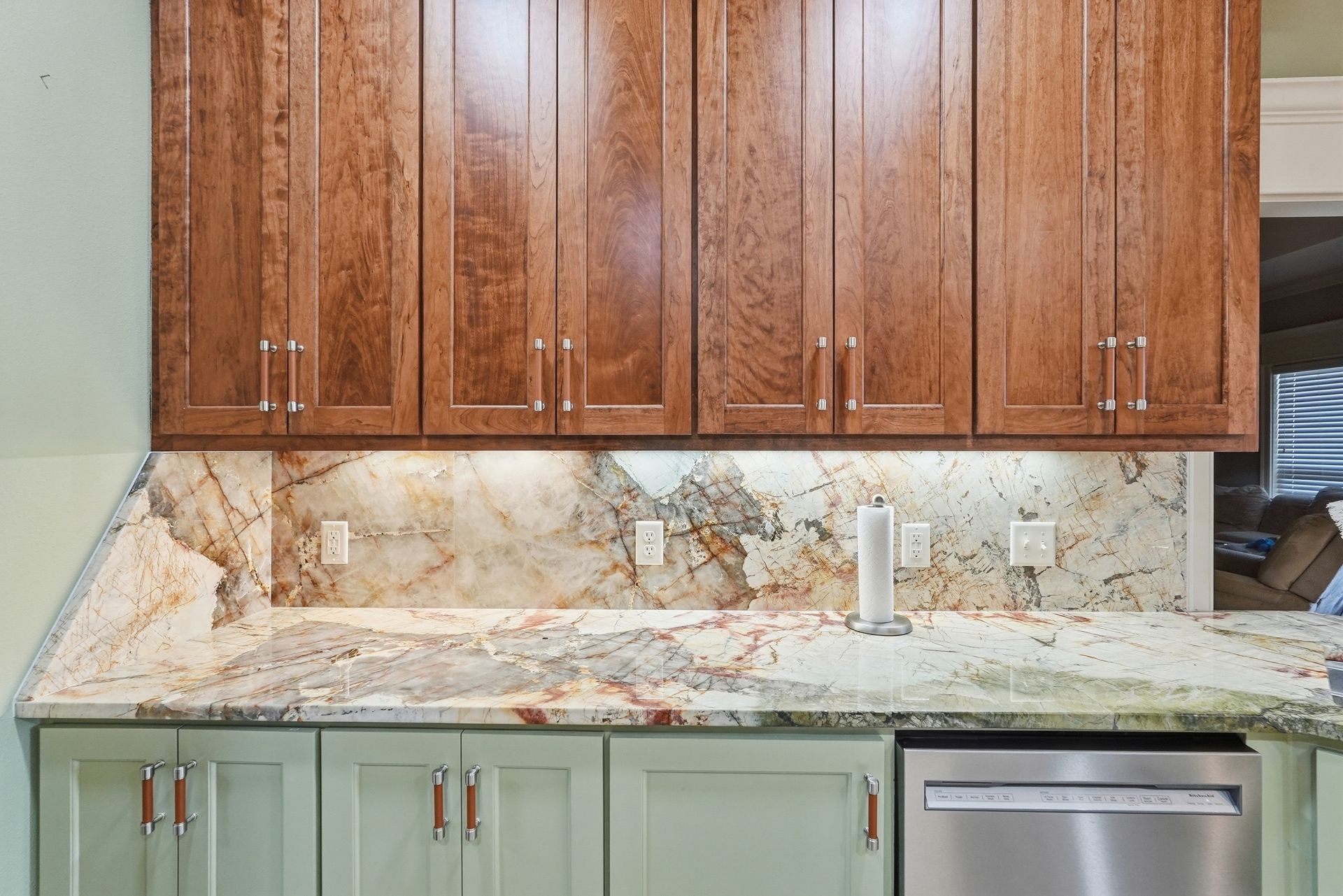 A kitchen counter with patterned stone backsplash, wood upper cabinets, light green lower cabinets, and a dishwasher.