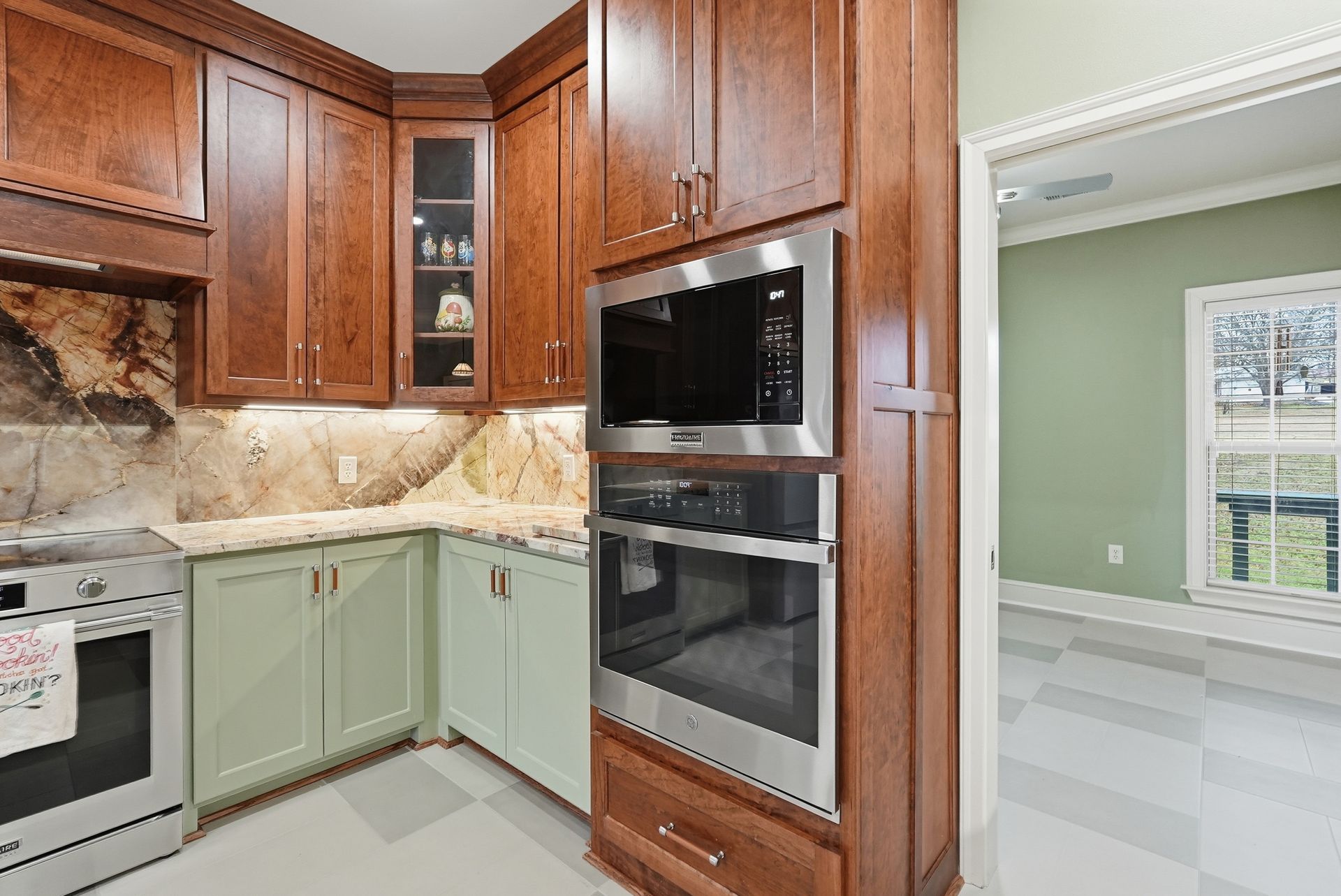 A kitchen with wooden upper cabinets, pale green lower cabinets, stainless steel appliances, and a marble backsplash.