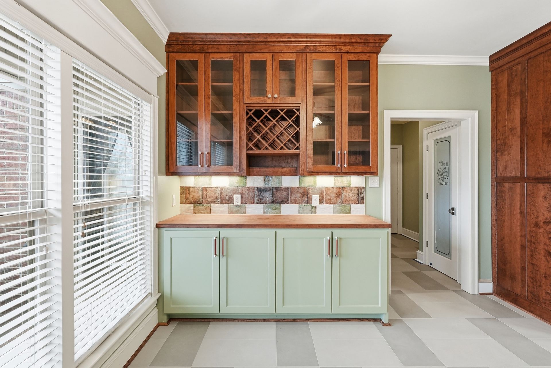 A home wet bar with light green cabinets, a wood-topped counter, a tiled backsplash, and glass-front wood upper cabinets.