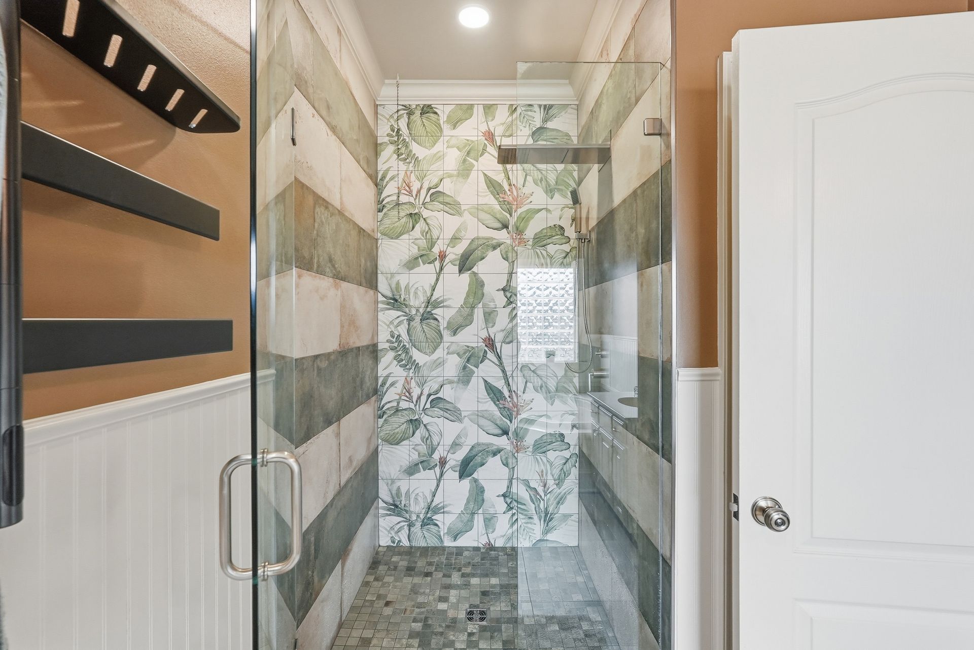A walk-in shower with a glass door, floral-patterned tiled back wall, and striped side walls with black shelving.