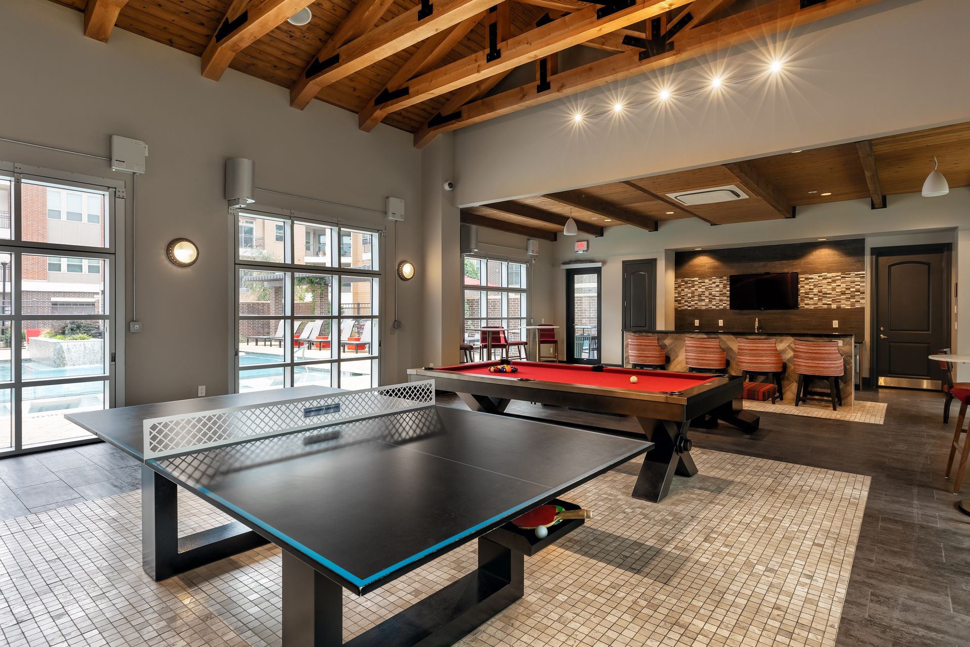Recreation room with a ping pong table, pool table, and seating, overlooking a pool area. Exposed wooden beams and neutral tones create a modern space.