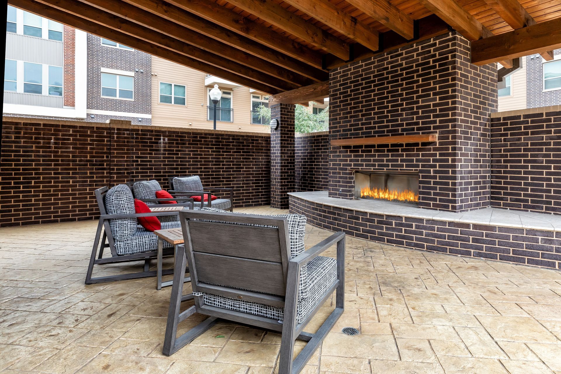 Outdoor seating area with brick fireplace and wooden ceiling. Chairs with patterned cushions surround a small table.