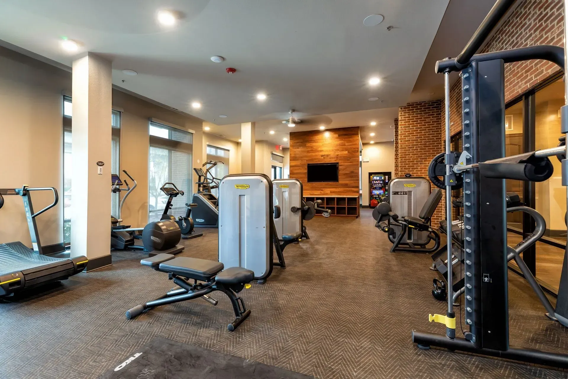 A modern gym with various exercise machines, a TV, and brick accents; the floor is covered in black rubber.