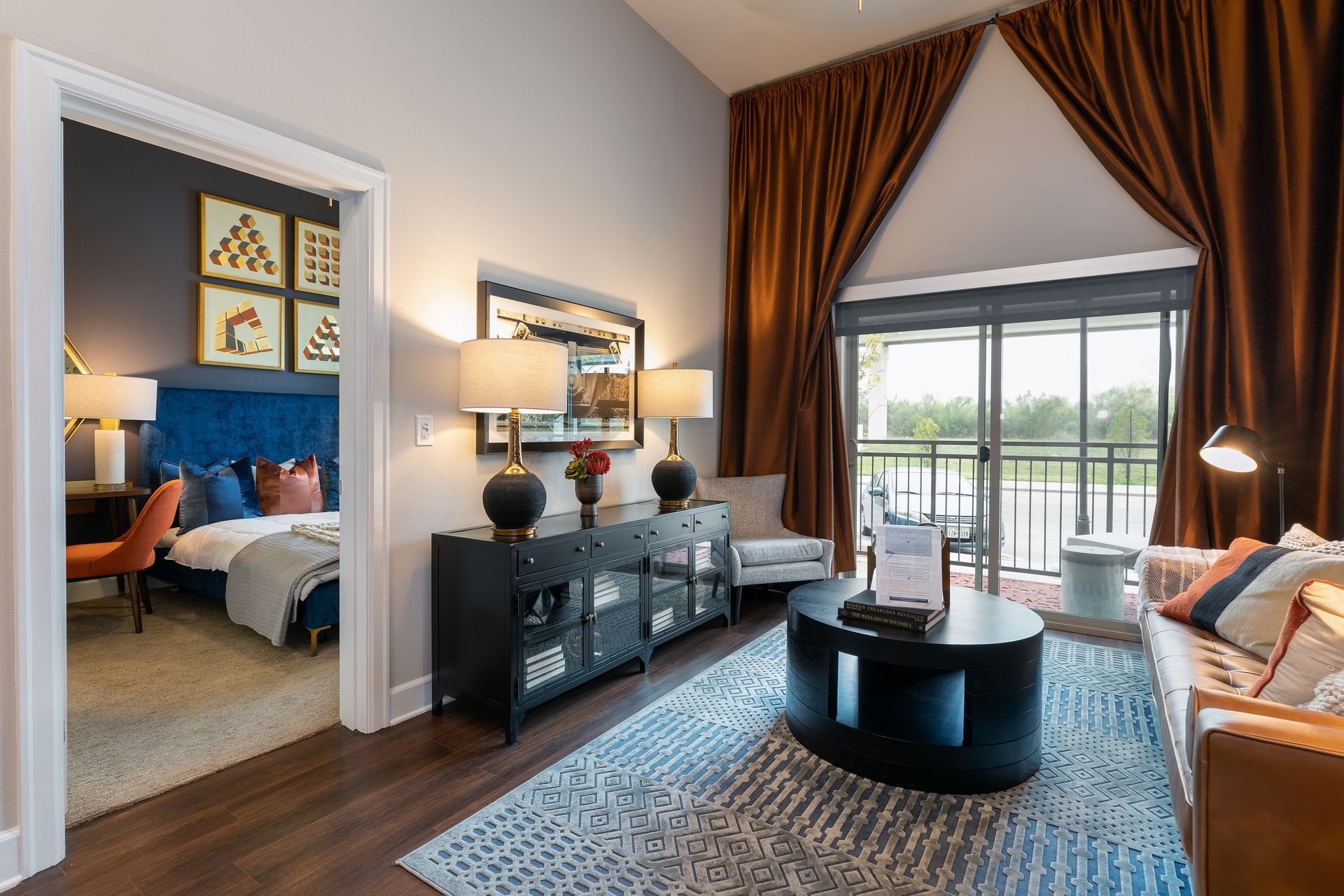 Interior view of a living room and bedroom; features a dark-colored couch, rug, and window curtains. The bedroom has a blue bed frame.
